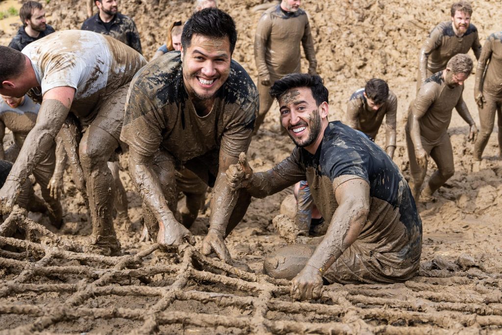 Twee jongens die lachend in de camera kijken en onder de modder zitten terwijl ze aan een net omhoog klimmen. Foto voor Modder uit je kleding krijgen na een obstacle run